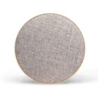 The image shows a round portable speaker with a fabric front and a thin golden rim, set against a plain, white background.