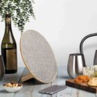 A tabletop scene with a bottle of wine, two stainless steel cups, a smartphone, a round speaker, and a platter of cheese, grapes, dried apricots, and crackers. A bowl of nuts and a hanging plant also adorn the wooden table.