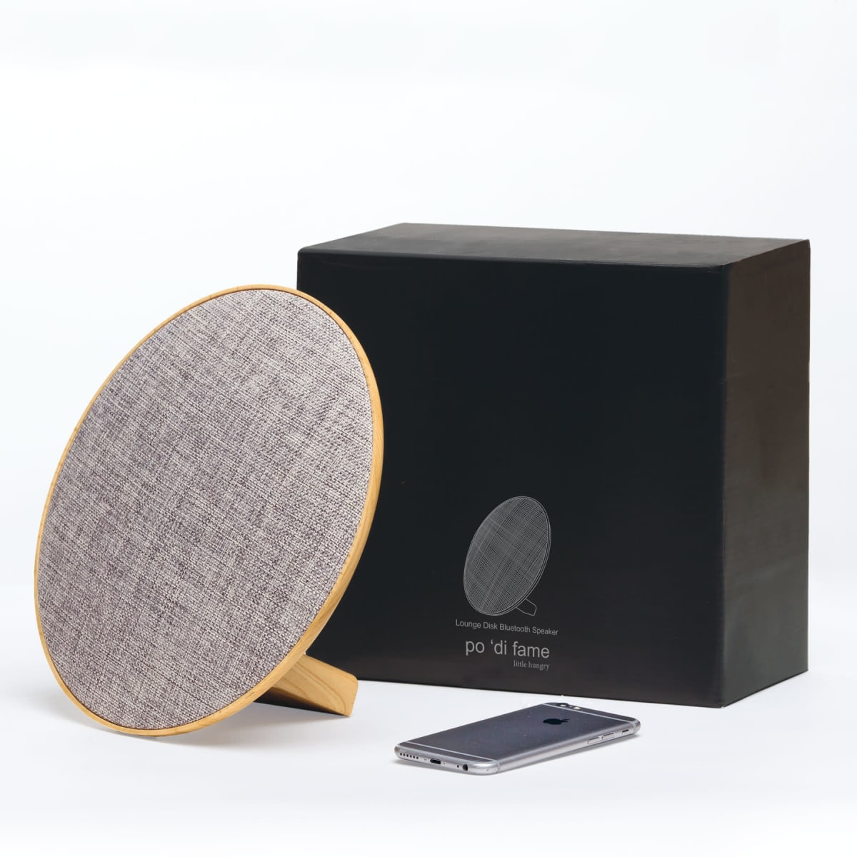 A modern, circular Bluetooth speaker with a fabric front and wooden frame, standing upright beside a black box. A smartphone lies flat on the surface nearby.