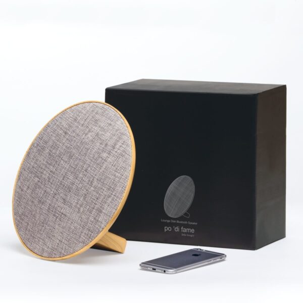 A modern round Bluetooth speaker with a wooden frame and fabric cover stands in front of its black packaging box with a minimalist design. A smartphone is placed nearby on a white surface.