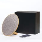 A round, fabric-covered wireless speaker with a wooden stand is placed beside a large, black rectangular box. A smartphone lies flat on a white surface in front of the speaker. The background is plain white.