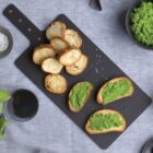 A slate cutting board with toasted baguette slices and green pea puree spread on some. Surrounding items include a bowl of flaky salt, fresh basil, two glasses of water, a bowl of green pea puree, and a small dish with mixed olives.