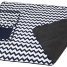 A folded picnic blanket with a black and white zigzag pattern. It has a black handle and a small pocket on one side, with a black waterproof backing visible underneath.
