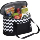 A black and white zigzag-patterned cooler bag is open, revealing its contents: a can of Coke, packets of cashews, a juice bottle, an apple, and a lemon. The bag has black handles and a front pocket.