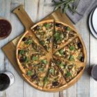 A vegetarian pizza topped with olives, mushrooms, and greens, sliced on a wooden board. Nearby are a bowl of tomato sauce, a bottle of red wine, a glass of wine, two white plates, and a gray napkin on a wooden table.