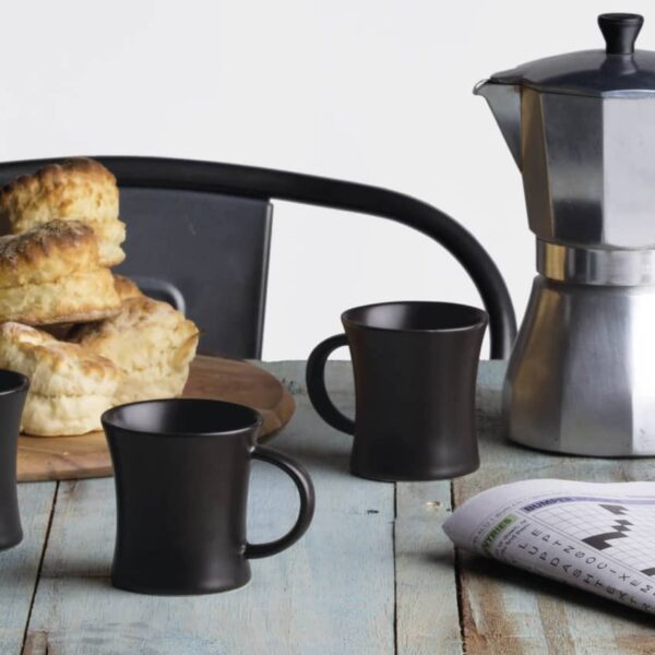 A rustic wooden table with black coffee mugs, a metallic stovetop coffee maker, a plate of scones, and a newspaper featuring a crossword puzzle.