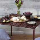 A small wooden table with a variety of cheeses on a round board, crackers, nuts, and grapes. Two glasses of red wine are placed on the table. A vase with flowers is in the center, and pillows are on the floor nearby.