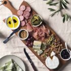 A wooden charcuterie board with salami, prosciutto, pickles, assorted cheeses, nuts, grapes, and a bowl of jam. Dishes of olive oil and olives, and breadsticks in a jar, are nearby on a table with a light tablecloth and leafy garnishes.