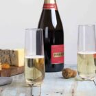 Two champagne flutes filled with bubbly champagne sit on a rustic wooden table. Behind them is a bottle of Piper Heidsieck champagne. A cheese board with blue cheese, crackers, and almonds is nearby. A cork rests on the table.