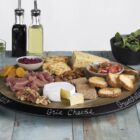 A charcuterie board with prosciutto, brie cheese, bruschetta, sliced bread, nuts, dried apricots, olives, salad, and cheese. Two glasses with lemon water, and bottles of oil and vinegar are nearby on a rustic table.