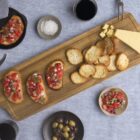 A wooden board with sliced bread, diced cheese, and bruschetta topped with diced tomatoes and onions. Nearby are small bowls of olives, two glasses of red wine, and salt. A knife is on the side, and a gray cloth is partially visible.