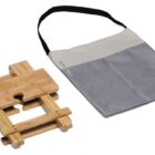 A foldable bamboo book stand with adjustable slats is placed next to a gray and beige carrying bag with a black strap. The book stand features a cutout design and natural wood finish.
