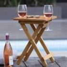 A small wooden folding table holds two glasses of rosé wine. An open wine bottle and cork are on the ground beside a cushion, all set near a poolside.