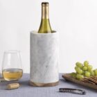 A marble wine cooler holds a bottle of wine, with two filled glasses nearby. A wooden tray displays green grapes, crackers, and cheese. A corkscrew and cork rest on a gray tablecloth, creating an elegant scene.