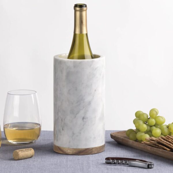 A marble wine cooler holds a wine bottle, flanked by two filled wine glasses. A wooden tray displays green grapes, crackers, and cheese. A corkscrew lies nearby on a gray tablecloth.