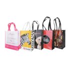 Five colorful tote bags with different designs stand upright. From left to right: pink with text, zebra photo, dog photo, scuba diver logo, and abstract design. Each bag has long handles.