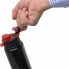 A hand wearing a blue checkered shirt holds a black water bottle with a red lid by its handle against a white background.