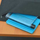 A black bag partially unzipped on a wooden table reveals a blue notebook and a clear water bottle inside.