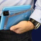 A person holds a clear, flat water bottle resembling a book in their arm, along with a blue notebook. They are wearing a light blue shirt and a black watch.