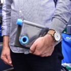 A person is holding a transparent, uniquely shaped water bottle with a blue cap. They are wearing a light blue button-up shirt and dark jeans, and a watch is visible on their wrist. The background includes shelves with various items.