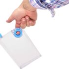 A person wearing a plaid shirt holds a transparent, rectangular water bottle with a blue lid and handle. The bottle is being held by the finger through the hole in the handle against a white background.