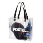 Clear tote bag with black handles containing a navy blue cap, a bottle of sunscreen, blue canister, and a black rectangular object. The word FOXTEL is printed in white on the bags front.