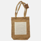 A brown jute tote bag with a white rectangular pocket on the front. It has two handles and a tag attached. The background is plain white.