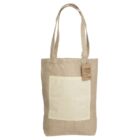 A beige jute tote bag with long handles and a large cream-colored pocket on the front. A small brown tag with text and a design hangs near the handle.