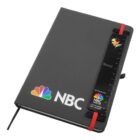 A black notebook with the NBC logo in color at the bottom. Its held closed by a red elastic band, and features a black ruler attached to the cover with text and a small image of a person on it.