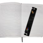 An open lined notebook with a black bookmark featuring a ruler, an image of a man, and NBC branding. The bookmark is placed vertically across the right page.