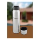A stainless steel thermos with a black lid and an additional detached lid used as a cup. The thermos has amazon.com printed on its side. The background is blurred, showing a hint of a building and greenery.
