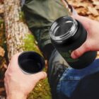 A person sitting outdoors holds a black thermos in both hands. One hand holds the thermos lid, which also functions as a cup. The background shows a fallen tree trunk and mossy ground, suggesting a natural setting.