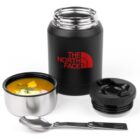 A black thermos with The North Face logo, a detachable lid, and a cup filled with soup, alongside a foldable spoon. The thermos has a sleek design, and the soup appears to have vegetable pieces and herbs.