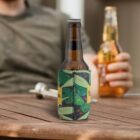 A person holding a beer bottle seated at a wooden table. In the foreground, another beer bottle is in a green and yellow tropical-themed koozie. The background is slightly blurry, focusing attention on the bottles.