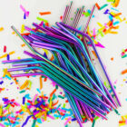 A pile of colorful metallic straws with bent tips lies on a white surface. Surrounding the straws are scattered pieces of vibrant, multicolored confetti.