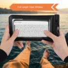 A person holding a waterproof pouch with a phone inside, displaying text, while sitting by a body of water. The pouch has a clear window. Feet are visible in the background, suggesting relaxation by the water. Orange banner reads Full Length Clear Window.