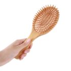 A hand holding a wooden hairbrush with an oval shape and wooden bristles, set against a plain white background.