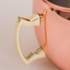 Close-up of a shiny copper mug featuring a gold-colored handle. The surface reflects light, highlighting its smooth texture. The mugs curved shape and metallic finish create an elegant and polished appearance.