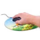 A hand using a black wired computer mouse on a vibrantly colored mouse pad featuring a landscape with green grass, flowers, and a small house under a blue sky.
