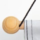 Close-up of a wooden dowel with a round end, partially wrapped in a white and gray striped fabric. A black cord is tied to the dowel, set against a plain white background.