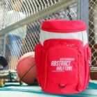 A vibrant red backpack with Background Abstract Halftone printed on it is placed on the ground near a basketball and skateboard. There is a chain-link fence in the background with a blurred figure behind it.