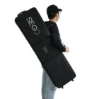 Person in a black cap carrying a large, rectangular black case with SEGO written on it over their shoulder. They are wearing jeans and a black top.