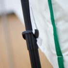 Close-up of a black metal pole with a locking mechanism attached to a white fabric with green trim. The fabric appears to be part of a larger structure or fixture, and the background is blurred.