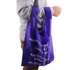 A person wearing a gray sweatshirt is holding a purple reusable shopping bag. The bag has white text on it, including contact information and business details. The person is positioned against a white background.