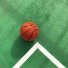 A basketball sits on a green court, positioned near the corner formed by white boundary lines. The sunlight casts a shadow of the ball on the textured surface.