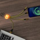 A glowing pumpkin-shaped keychain with multiple charging cables is plugged into a laptop and an iPhone. The phone screen displays a partially charged battery symbol. The setup is on a wooden surface.