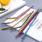A collection of colorful metal straws in golden, silver, blue, and purple hues are placed on an open book on a gray surface. Lemons and coriander are partially visible in the corner.