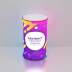 A cylindrical object with a colorful abstract geometric design. The surface features purple, yellow, pink, and white shapes with the text Abstract Geometric Colorful Background in the center. It stands on a light gray surface.