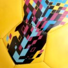 Close-up of a yellow soccer ball featuring a vibrant, geometric pattern. The design includes black, pink, blue, and light blue pixels forming a dynamic, abstract shape across the hexagonal panels.