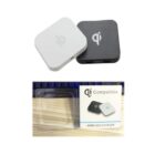 Two square wireless chargers, one white and one black, each with a Qi symbol. Below, a product package displays the chargers with Qi Compatible Wireless Charger labeled. Packaging is transparent, showing a wooden texture underneath.